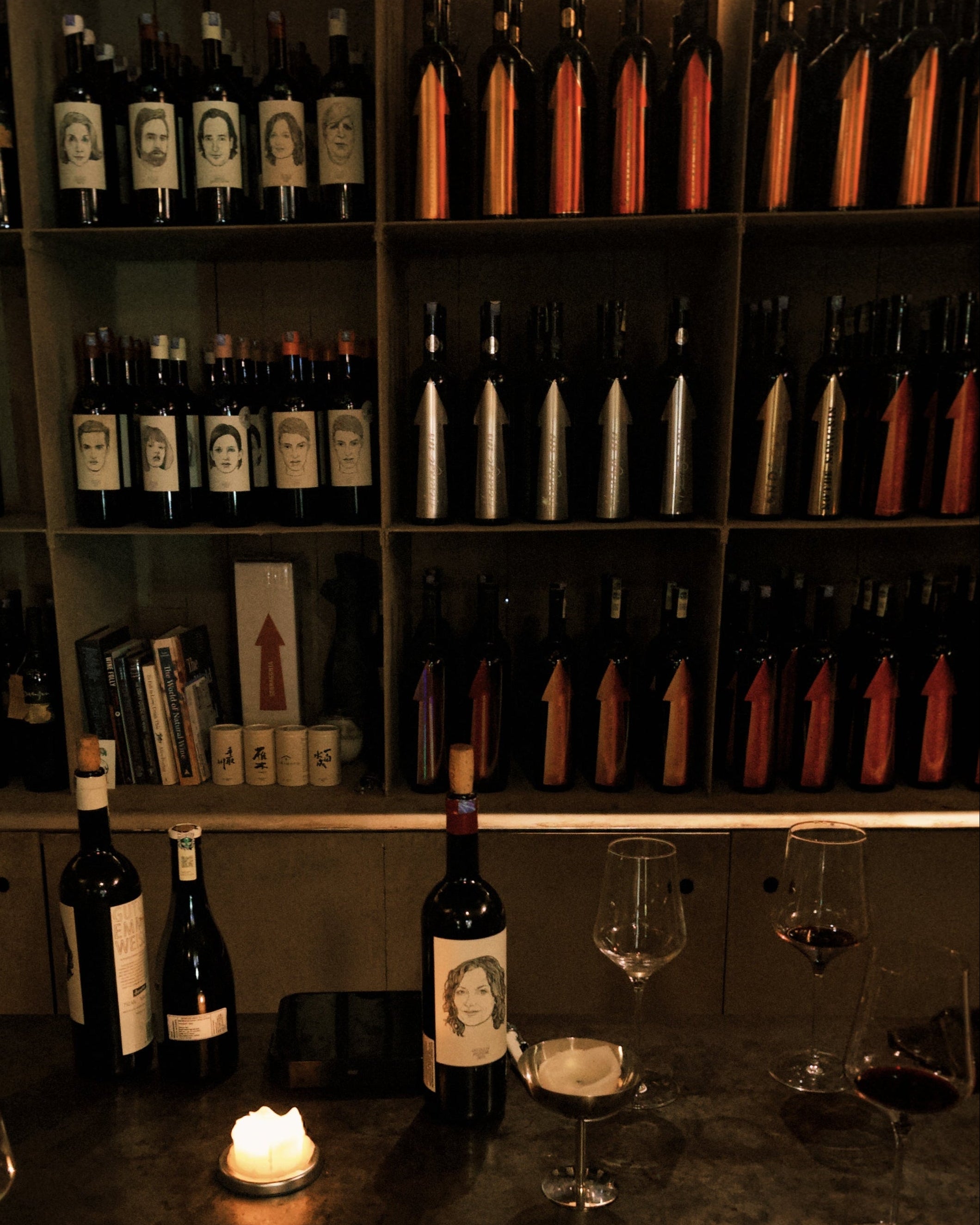 Wine bottles and glasses on a bar with a candle, in a dimly lit setting.